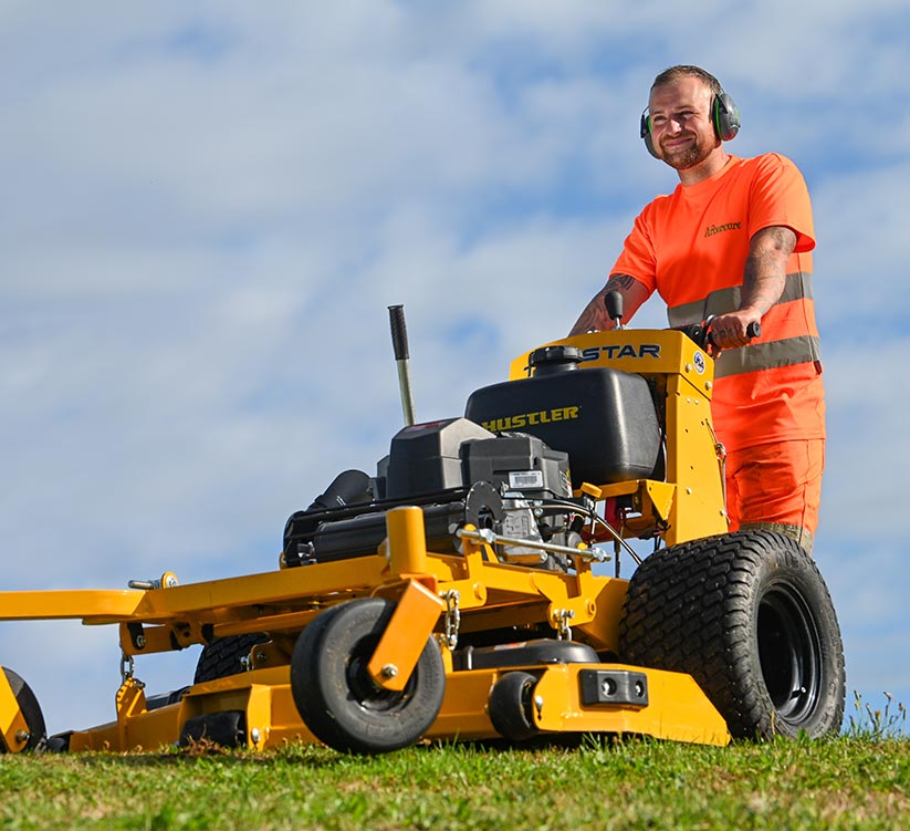 Arborcure grounds maintenance employee in orange high visibility uniform mowing grass with industrial law mower